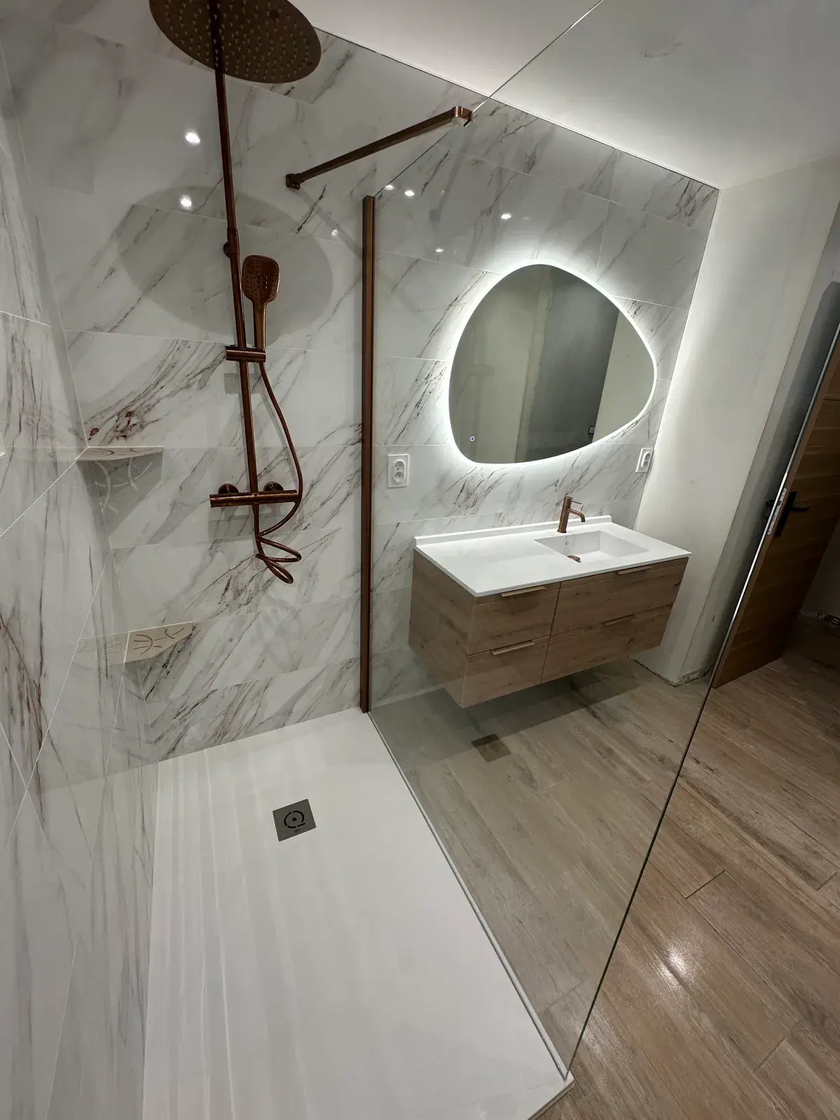 Salle de bain réalisée par NC Plomberie Chauffage : douche à l'italienne en marbre blanc, colonne de douche cuivrée, miroir galet rétroéclairé et meuble vasque suspendu en bois