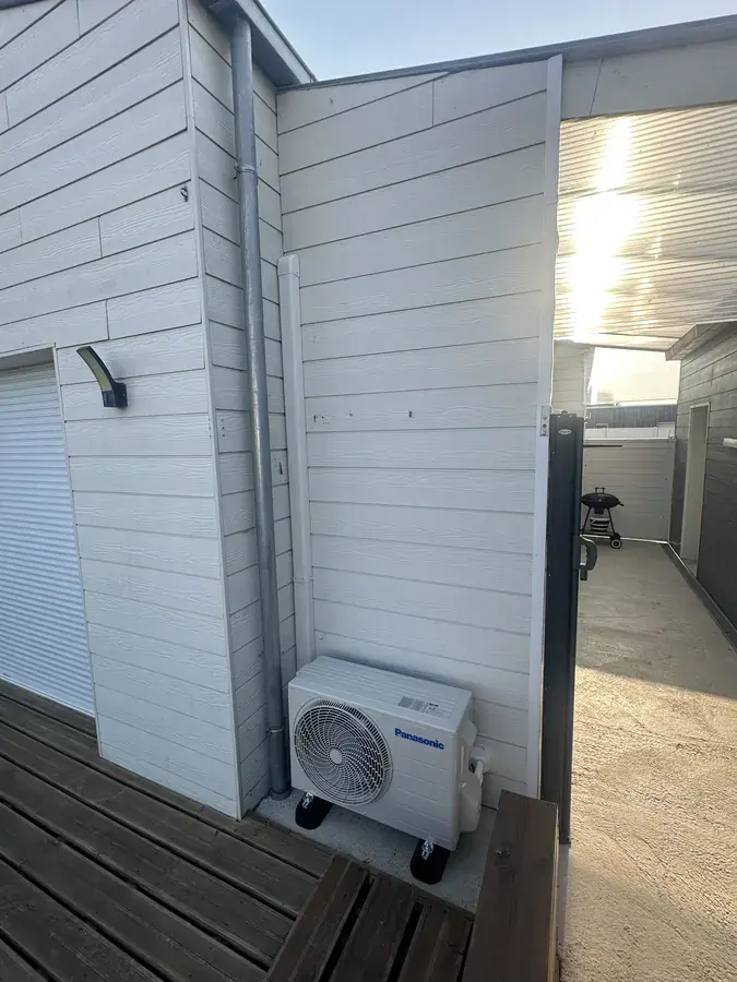 Unité extérieure de climatisation posée sur une terrasse bois au pied d'un bardage blanc